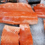 Several pieces of sushi grade salmon fillets are displayed on crushed ice. The salmon has a vibrant orange color with visible white marbling, showcasing its exceptional quality and freshness.