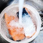 A piece of salmon being rinsed under running water in a metal sink, key to preparing salmon. The water creates bubbles on the fish's surface as it fills the bowl surrounding the vibrant fillet.