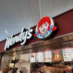 A Wendy's fast-food outlet with its iconic logo featuring a red-haired girl. The menu displays items like the "Hot Honey" burger. Customers are seen at the counter under a bright ceiling.