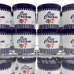 A display freezer filled with tubs of frozen ice cream. Each tub has a navy blue lid and a label with "Ice Cream" in bold letters, striped design, and images of three ice cream cones. The tubs are safely arranged on two shelves.