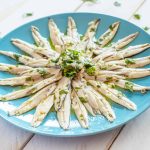 A blue plate artistically arranged with salty marinated anchovies in a circular pattern, garnished with chopped parsley, on a light wooden table.