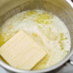 A stick of butter is melting in a saucepan on a stovetop, reaching its boiling point. The butter is partially melted and frothy, with some solid portions still visible, creating a mixture of liquid and bubbles.