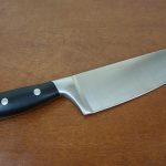 A sleek stainless steel kitchen knife with a black handle, accented by three silver rivets, rests elegantly on a wooden surface, showcasing its expertly forged blade.