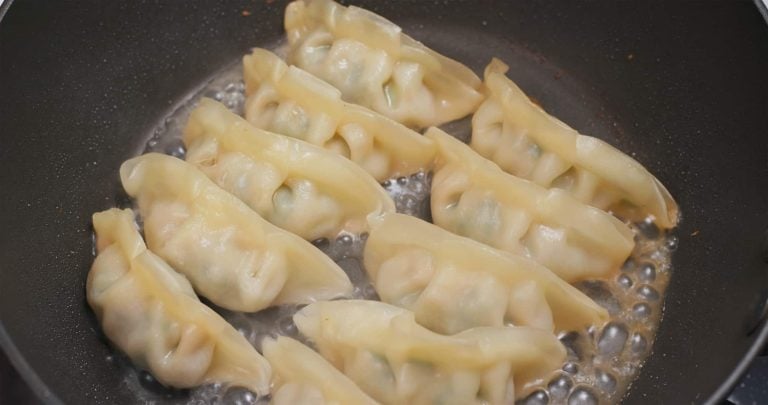 Eight dumplings sizzle in a black skillet, with oil bubbling around them, showcasing a light golden-brown edge. Whether you savor them hot or eat cold, these dumplings promise mouthwatering delight.