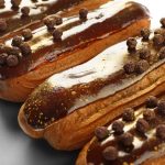 Close-up of several eclairs topped with glossy chocolate glaze and sprinkled with round chocolate pieces. The pastries, boasting a creamy filling within their delicate shells, are arranged in a neat row on a reflective surface.