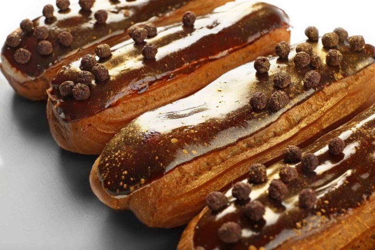 Close-up of several eclairs topped with glossy chocolate glaze and sprinkled with round chocolate pieces. The pastries, boasting a creamy filling within their delicate shells, are arranged in a neat row on a reflective surface.