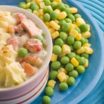 A serving of delectable fish pie topped with creamy mashed potatoes in a ramekin, accompanied by sides of mixed peas and corn on a blue plate. A fork with a polka dot handle rests elegantly on the plate, perfect for pairing with your favorite wine.