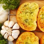 Vegan garlic bread with a golden crust sits on a cooling rack, surrounded by fresh parsley and whole garlic bulbs on a wooden cutting board. The bread is generously topped with herbs and garlic butter.