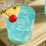 A refreshing blue cocktail served in a glass filled with ice, almost as cold as a freeze. It is garnished with a pineapple slice and a red cherry, placed on a wooden board, reminiscent of the tropical vibes of Hawaiian Punch.