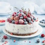 A frosty cheesecake topped with a mix of frozen berries, including raspberries, blueberries, and cherries, sits on a white plate. As you ponder how long it'll take to thaw, the background features a light blue surface scattered with berries, while a spoon rests nearby.