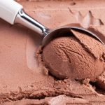 A metal ice cream scoop is lifting a smooth, round scoop of chocolate ice cream from a container. The creamy texture is perfect, thanks to the storage in the freezer, and the scoop's handle is white.