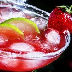 Close-up of a vibrant red cocktail in a glass with a sugared rim, creatively garnished like a Margarita with ice cubes, lime wedges, and a fresh strawberry. The drink's surface glistens against a dark background, creating a refreshing, summery appearance.