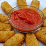 A circular arrangement of golden-brown fried mozzarella sticks surrounds a small glass bowl of rich red marinara sauce on a white plate. Serve with mozzarella sticks for the perfect crispy and savory treat.