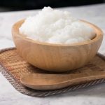 A wooden bowl filled with safe-to-eat mushy rice porridge sits on a square woven mat. A wooden spoon rests beside the bowl, all placed on a marble surface.