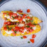 Two grilled corn cobs on a plate, topped with sour cream, diced tomatoes, and chopped herbsāan irresistible combination of fresh ingredients similar to pico de gallo.