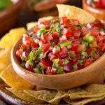 A bowl of fresh Pico de Gallo, bursting with chopped tomatoes, bell peppers, onions, and cilantro, sits invitingly in a clay bowl surrounded by tortilla chips. The vibrant mix of ingredients captures the eye, while limes and additional bowls are artfully blurred in the background.
