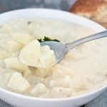A bowl of creamy potato soup with chunks of potato, garnished with parsley. A spoonful is lifted from the bowl, a perfect guide to savor the flavors. In the background, a piece of bread rests just like it would in your fridge, waiting to accompany this comforting delight.