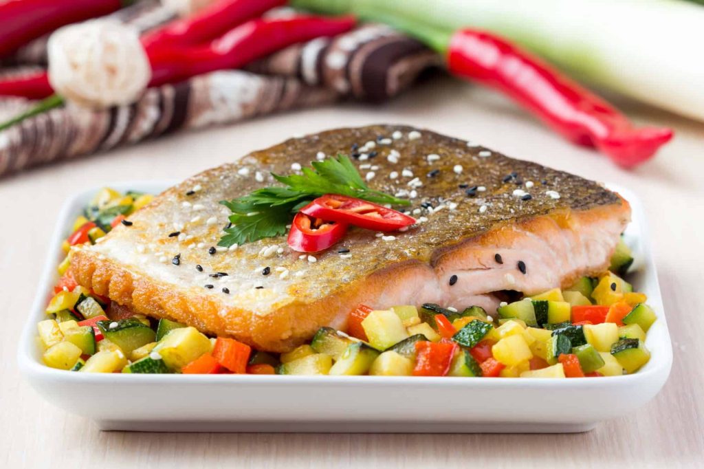 A crispy salmon fillet, with perfectly crackled salmon skin, is garnished with sliced red chili and parsley and sits on a bed of colorful diced vegetables, including zucchini and bell peppers. Red chili peppers and leeks are visible in the background.