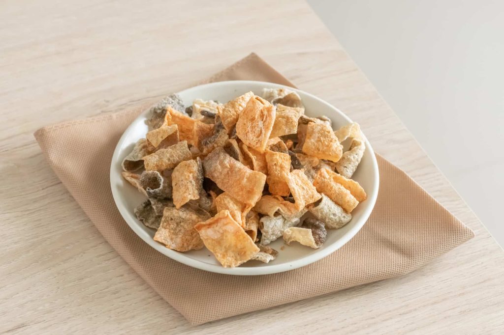 A white plate with mixed crispy pork snacks, including pork rinds, cracklings, and salmon skin, sits on a light brown napkin on a wooden table.