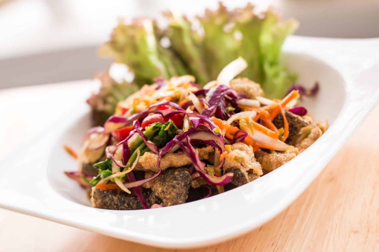 A vibrant dish featuring crispy fried salmon skin topped with a colorful mix of shredded cabbage, carrots, and red onions. Garnished with fresh lettuce leaves, it's presented on a white plate set on a wooden surface.