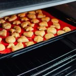 A baking tray filled with small, golden-brown, cookie-shaped pastries sits safely on the middle rack of an open oven. The treats rest on a red silicone mat, perfectly designed for their delightful transformation. The oven door remains slightly ajar.