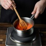 A person wearing a black apron skillfully stirs red tomato sauce with a wooden spoon in a small saucepan on the stove, showcasing expert cooking techniques. The scene unfolds in a cozy kitchen, where simmering tips like using a lid to maintain flavor are put into practice on the wooden countertop.