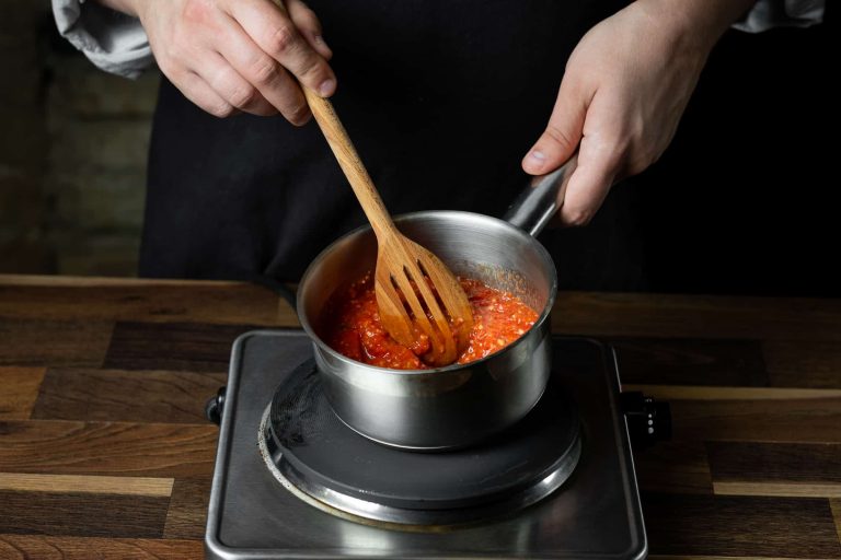 A person wearing a black apron skillfully stirs red tomato sauce with a wooden spoon in a small saucepan on the stove, showcasing expert cooking techniques. The scene unfolds in a cozy kitchen, where simmering tips like using a lid to maintain flavor are put into practice on the wooden countertop.