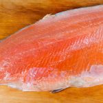 A fresh salmon fillet, ideal for making sushi, rests on a wooden cutting board, showcasing its vibrant orange flesh and smooth texture.