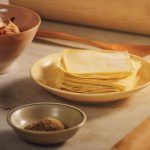 A plate of stacked wonton wrappers is centered on a kitchen surface, hinting at delicious creations to come. Nearby, a bowl contains shredded ingredients, and a small dish holds a brown spice. For optimal freshness, consider fridge storage to keep those wrappers ready for your culinary adventures.