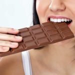 A person with dark hair is about to enjoy a large milk chocolate bar, eagerly holding it in their right hand. The background is a plain light color, emphasizing their moment of indulgence even amid thoughts of the paleo diet where chocolate isn't typically allowed.