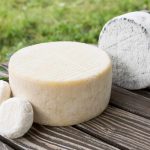 A variety of cheeses, including creamy goat cheese, are displayed on a wooden table outdoors. There is a large round cheese wheel and several smaller rounds, set against a rustic green background of grass.