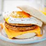 Breakfast sandwich with a runny fried egg, Quorn sausage patty, and melted cheddar cheese on a bun, placed on a white plate. A glass of orange juice is in the background.