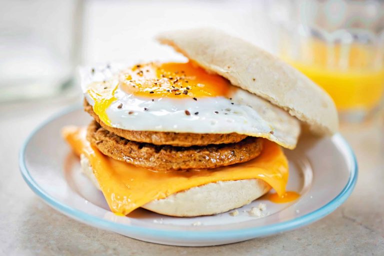 Breakfast sandwich with a runny fried egg, Quorn sausage patty, and melted cheddar cheese on a bun, placed on a white plate. A glass of orange juice is in the background.