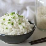 A black bowl of fluffy, perfectly cooked white rice garnished with chopped green herbs is the centerpiece of this image. In the background, a glass jar filled with uncooked rice and a pair of wooden chopsticks complement the minimalistic and clean presentation. The soft lighting and neutral tones enhance the simplicity and appeal of the dish.