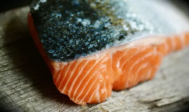 A close-up of a raw salmon fillet with skin graces a wooden surface. The vibrant orange flesh is marbled, illustrating the texture and freshness of the fish, while its silvery salmon skin adds an intriguing, textured contrast.