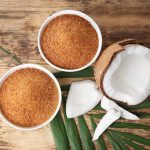 Two bowls of golden brown coconut sugar sit on a wooden surface next to a split coconut and green leaves, creating a stunning contrast with the white flesh. A staple in many Paleo recipes, this rustic scene invites you to explore why or why not to incorporate it into your diet.
