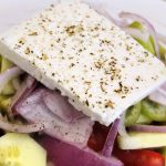 A fresh, paleo-friendly salad with cucumbers, tomatoes, purple onions, olives, and green peppers, topped with a thick slice of herb-sprinkled feta cheese in a clear bowl.
