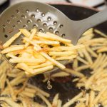 A used frying ladle gently lifts freshly fried shoestring fries above a simmering pan of oil, with more fries sizzling to perfection in the background.