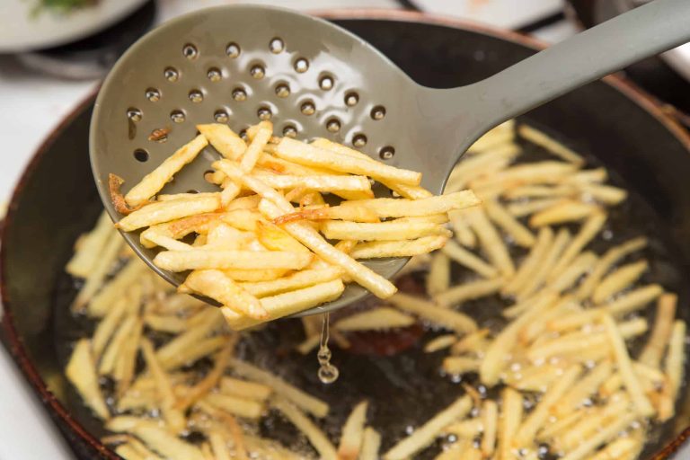 A used frying ladle gently lifts freshly fried shoestring fries above a simmering pan of oil, with more fries sizzling to perfection in the background.