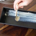 A hand uses a brush to spread butter on the inside of a rectangular metal baking pan placed on a wooden surface, ensuring it is thoroughly greased like one would with silicone baking pans.