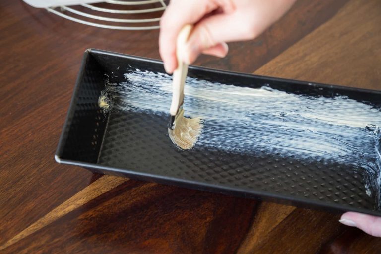 A hand uses a brush to spread butter on the inside of a rectangular metal baking pan placed on a wooden surface, ensuring it is thoroughly greased like one would with silicone baking pans.