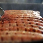 Close-up of sausages cooking on a grill with perfect grill marks. A pair of tongs is poised to turn them, as steam rises, indicating they're being cooked at ideal hot dog temperatures.