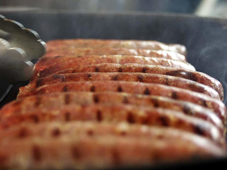 Close-up of sausages cooking on a grill with perfect grill marks. A pair of tongs is poised to turn them, as steam rises, indicating they're being cooked at ideal hot dog temperatures.