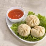 A plate with three steamed momos rests on a bed of green lettuce. A small white bowl filled with red chili sauce sits beside the dumplings on a light textured surface, offering a healthy and flavorful experience.