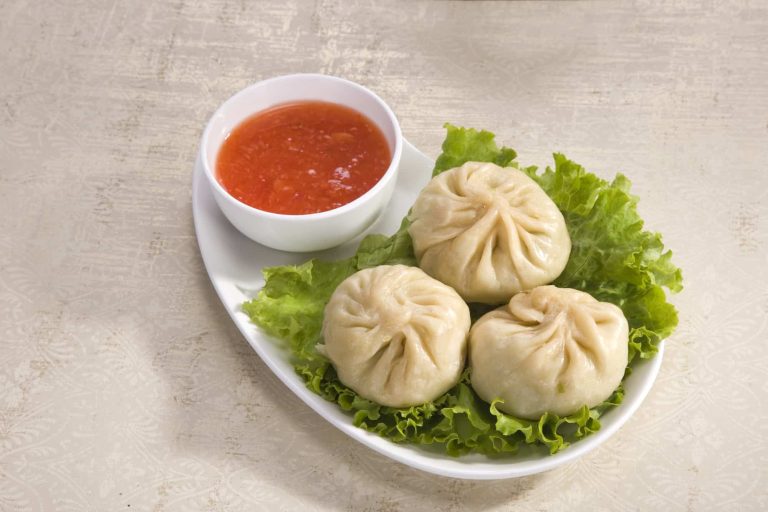 A plate with three steamed momos rests on a bed of green lettuce. A small white bowl filled with red chili sauce sits beside the dumplings on a light textured surface, offering a healthy and flavorful experience.