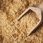 Close-up of nutritional yeast flakes with a wooden scoop partially buried in the tan, flaky texture. Loved by vegans for good reasons, this nutrient-rich ingredient brings a subtle complexity to any dish.