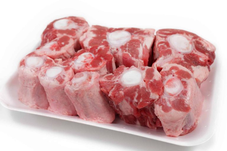 A white tray filled with raw oxtail pieces, displaying a mix of red meat and white bone sections. The oxtails are arranged closely together, showcasing their marbled texture and circular bone centers, perfect for cooking oxtails to perfection.