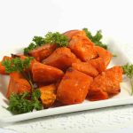 A white square plate with roasted sweet potato chunks, infused with a hint of orange-chipotle, garnished with fresh parsley. The plate is set on a decorative white doily.