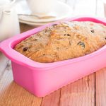 A freshly baked loaf of bread studded with dark fruit sits invitingly in a pink silicone mold on a wooden table. A white teapot and cup are blurred in the background, adding a cozy touch to this delightful step-by-step creation.