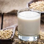 A glass of vegan milk sits on a wooden table, surrounded by soybeans. A wooden spoon holds more soybeans on the left. In the background, a bowl brimming with soybeans is set against a rustic fabric backdrop, offering a serene scene of plant-based milk tranquility.